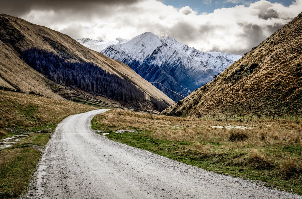 A winding gravel road through New Zealand's picturesque mountains under a cloudy sky.