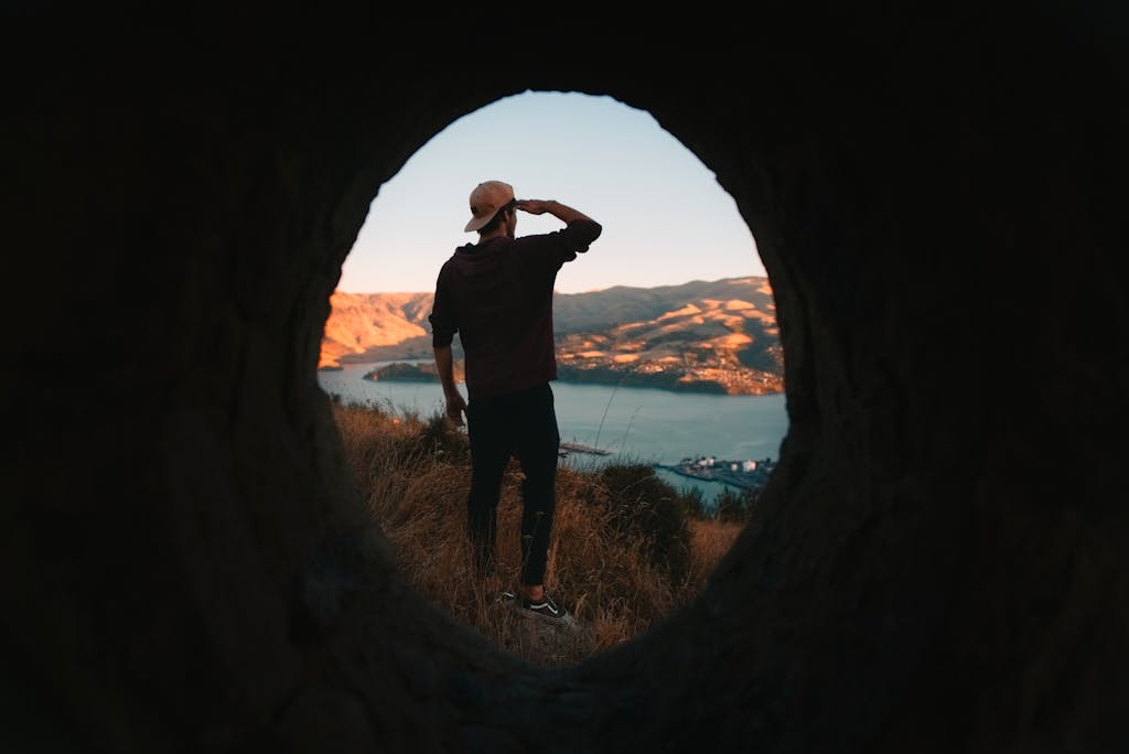 Man in a baseball cap observes a scenic coastal landscape through a tunnel at sunset.