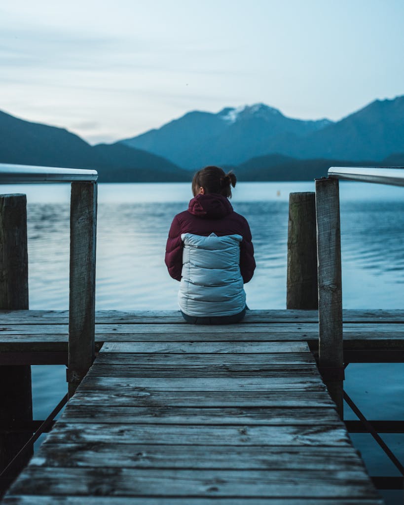 Woman sitting on wooden dock, enjoying the serene view of Lake Te Anau at twilight.